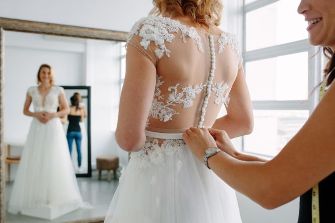 Bride standing in front of mirror and fitting dress in atelier with wedding assistant