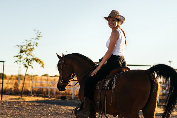 Young woman in hat riding a horse in the countryside at sunset