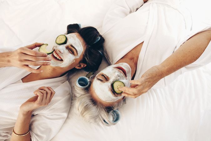 Two women in bathrobes lying on bed with face cream and cucumber slices on their eyes
