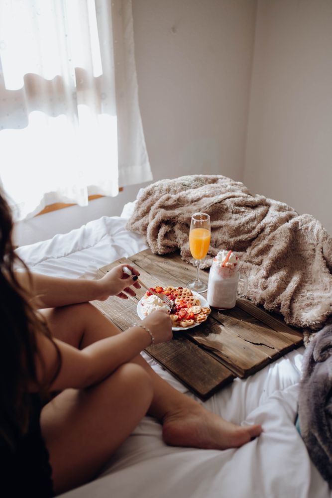 Young woman eating appetizing waffles and tasty drinks on tray on bed