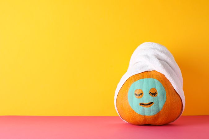 Pumpkin with facial mask and towel against yellow background, copy space