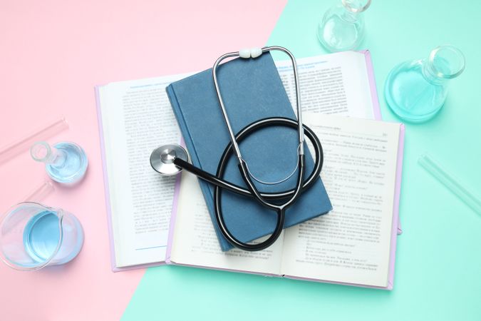 Books, stethoscope and flasks on pink and green background, top view