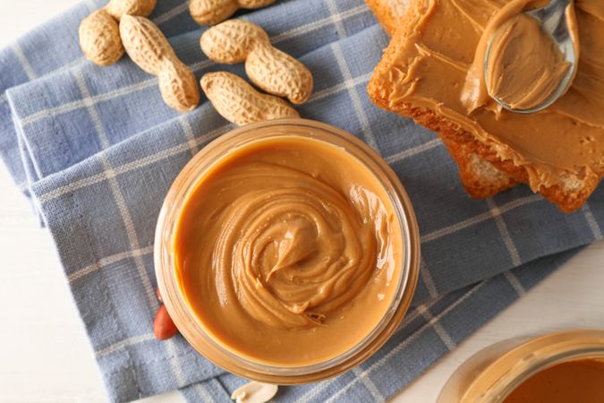 Glass jar with peanut butter, peanut, kitchen towel, spoon and peanut butter sandwich on wooden background, top view and closeup
