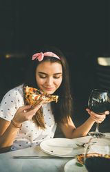 Young woman with red wine in hand, eating pizza outside in the sun, copy space