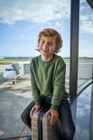 Positive kid smiling while sitting on luggage near window in air