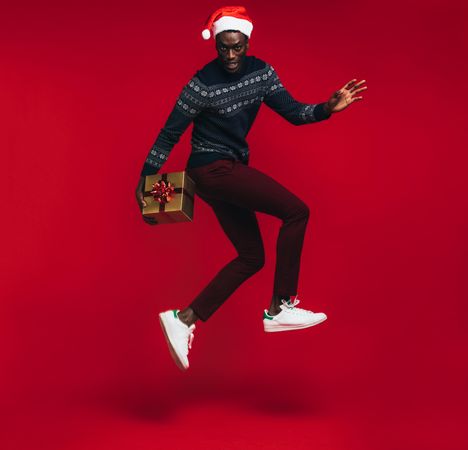 Young Black man wearing Santa hat jumping with gift box over red background