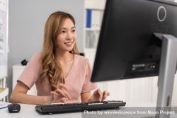 Happy Asian woman sitting in office and typing on keyboard while looking at monitor 5zq6P0