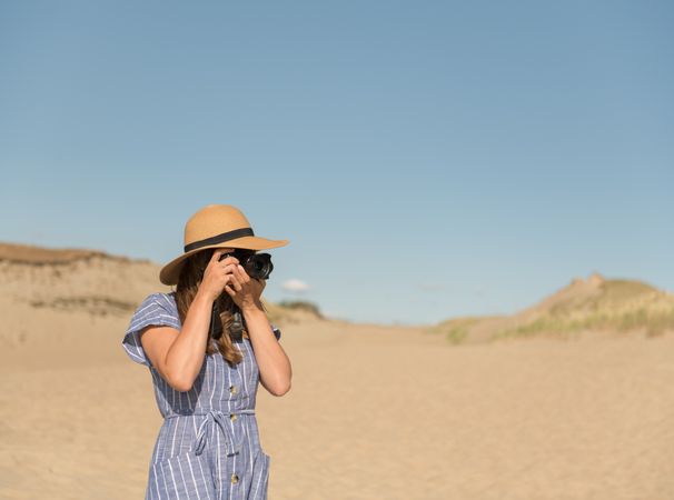 Back view of adult woman in straw hat and dress with camera taking photo on sand dune