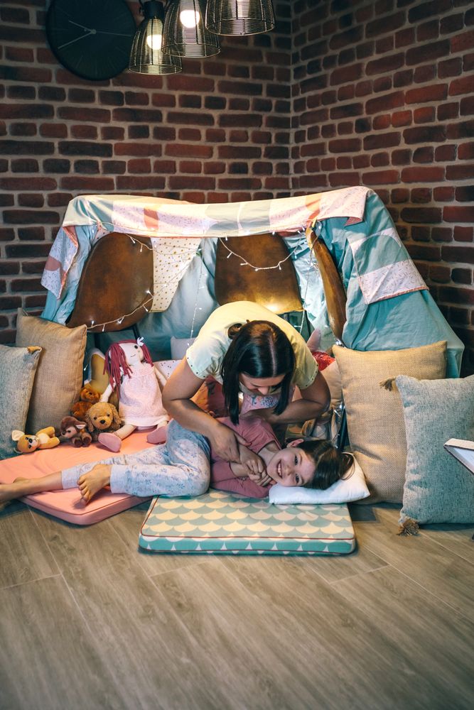 Woman tickling to little girl while laughing and shouting lying over cozy mats in night pajama party