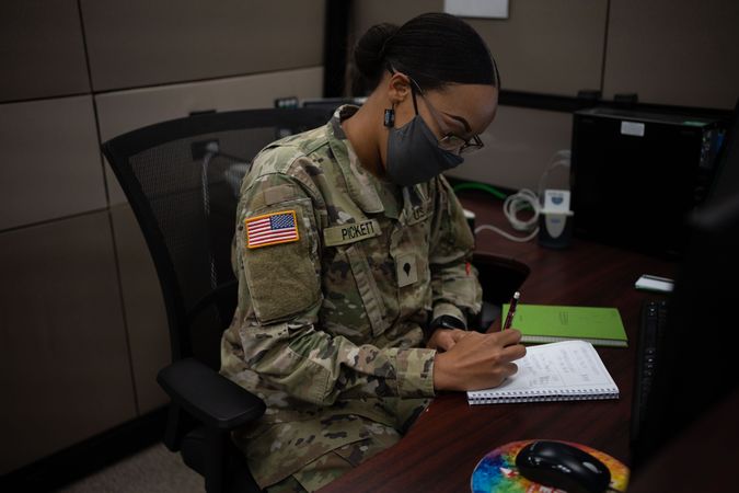 US female soldier with facemask sitting at her desk writing on notebook