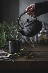 Woman pouring green tea in mug on wooden table with green herb