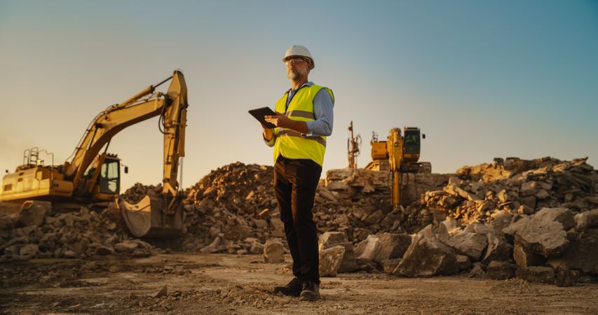 Male Real Estate Investor Wearing Protective Goggles And Using Tablet On Construction Site On A Sunny Day. Man Inspecting Building Progress. Excavator Loading Materials Into Industrial Truck