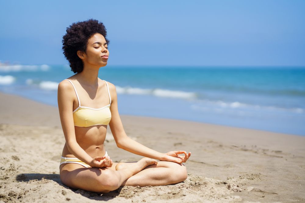 Woman in yellow swimwear sitting cross legged and meditating on sandy beach