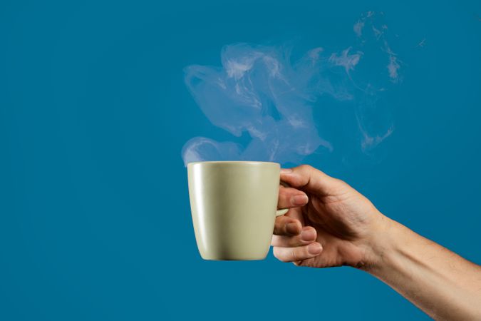 Hand holding a steaming hot coffee mug on a solid blue background.