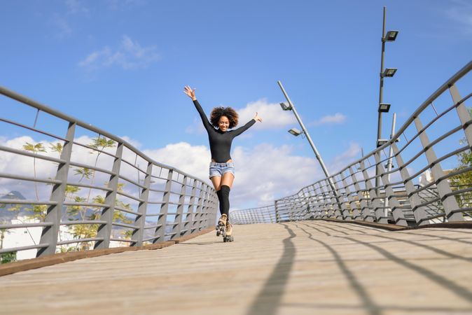 Woman with afro hairstyle in roller skates on wooden bridge with arms up