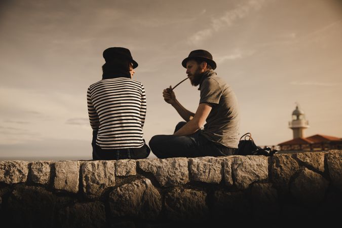 Man with pipe near woman with book on stones near lighthouse