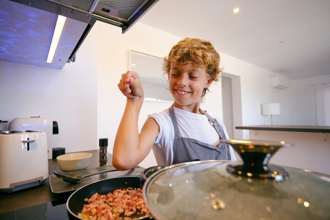 Satisfied boy salting bacon in frying pan while cooking
