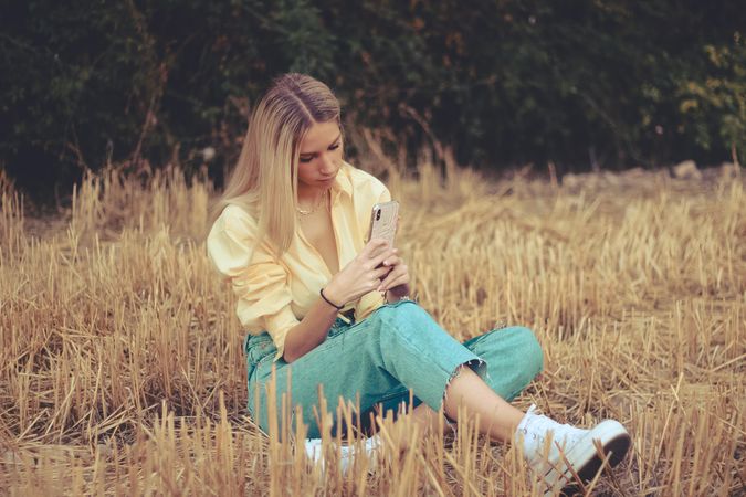 Blonde woman using smartphone sitting on brown grass