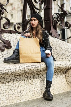 Woman in winter hat sitting outdoors holding a take away coffee and shopping bag