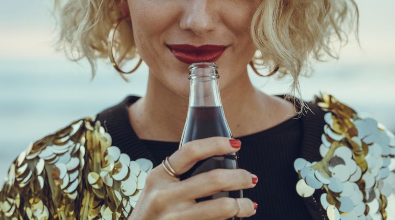 Cropped shot of female in sequined dress drinking cola at the beach