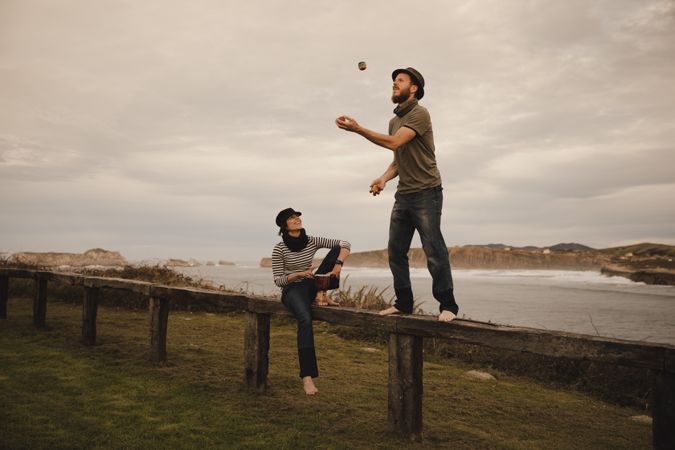 Man juggling balls on bench near woman with drum on shore