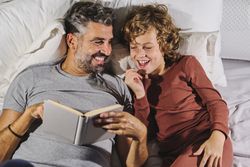 Laughing man and boy reading fairytale in bedroom