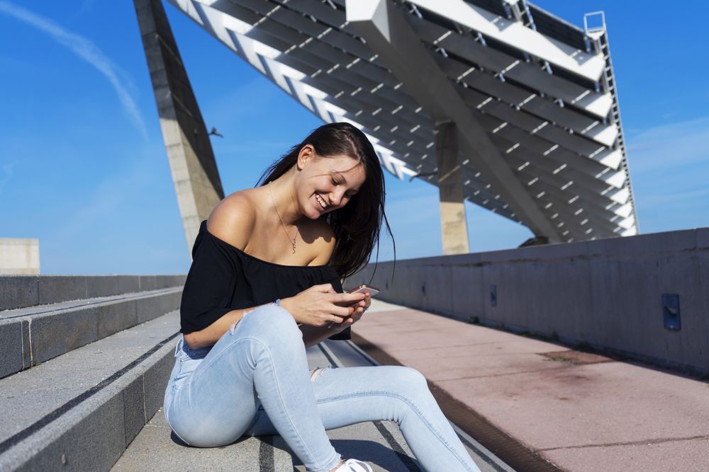 Young smiling woman with long hair sitting on promenade against harbor and checking phone
