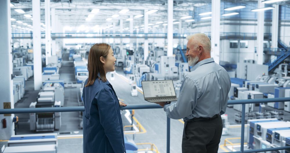 Mature Engineer and Young Japanese Technician Having a Conversation and Using Laptop Computer. Specialists Monitoring, Analyzing Conditions at a Modern Electronics Factory with Automated Robots