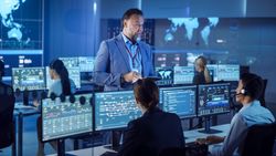 Bearded Manager Oversees Diverse Team in High-tech Control Room. Analysts Monitor Real-time Data on Multiple Screens.