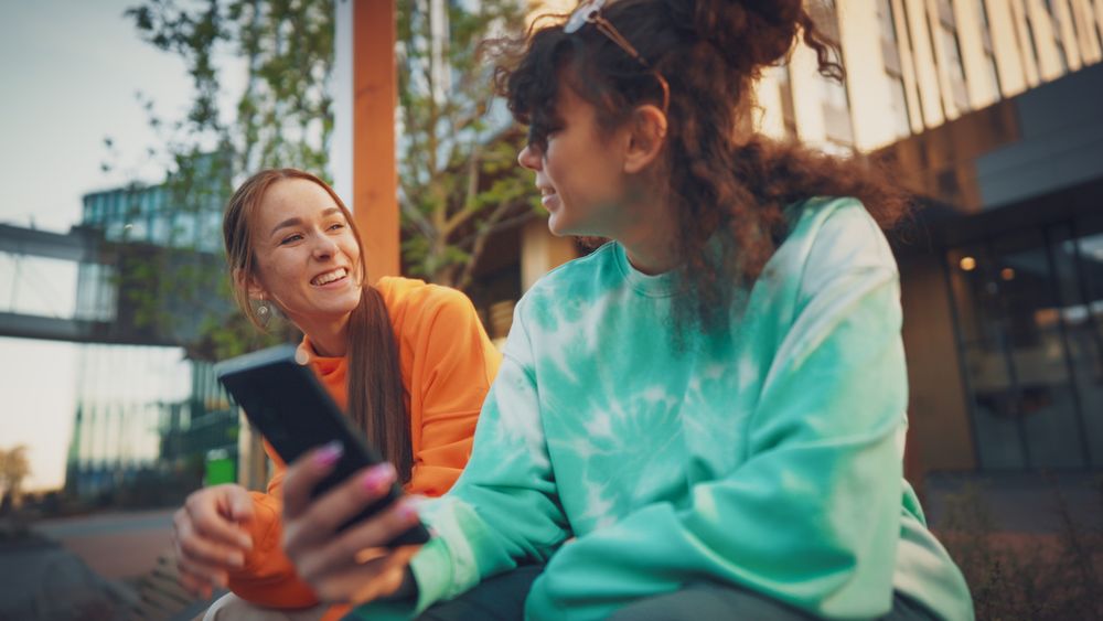 Two Young Women Sharing A Joyful Moment Outdoors, One Holding A Smartphone
