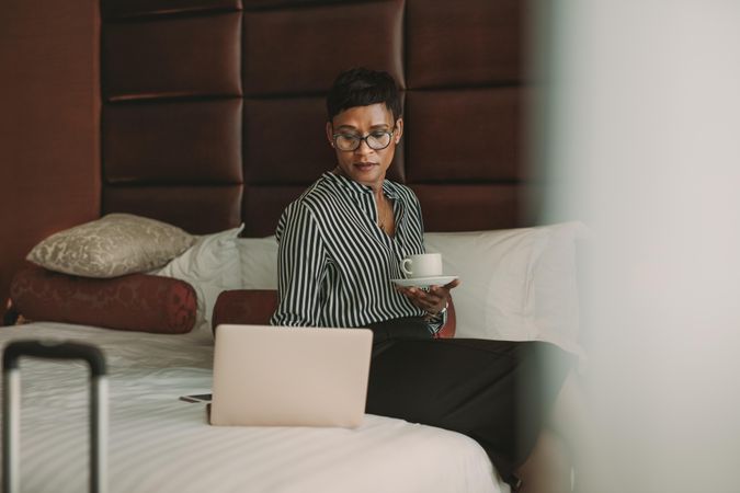Woman in formal clothes sitting on bed with coffee in hotel room using laptop