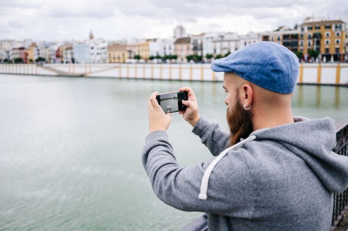 Guy on vacation in casual clothing making photo of bay on smartphone at quay
