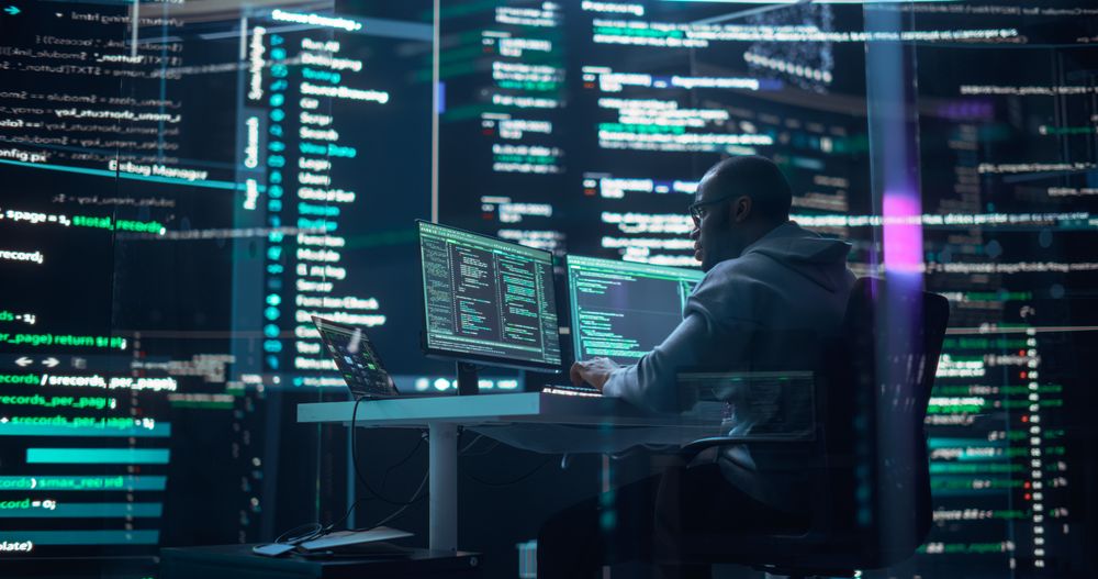 Black Male Developer Thinking and Typing on Computer, Surrounded by Big Screens Showing Coding Language.