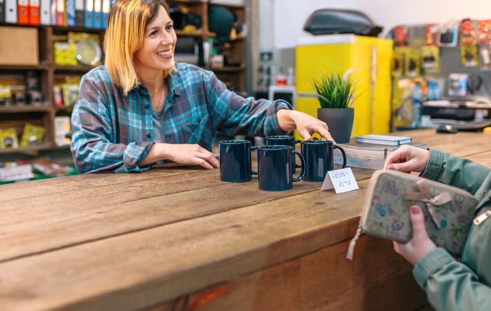 Smiling blonde woman employee with freckles showing blue mugs to customer in vintage shop