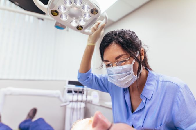 Female dentist wearing mask adjusting light with patient on chair at dental clinic