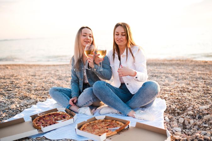 Two women eating pizzas and drinking wine sitting on sand beach