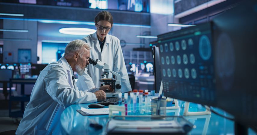 White Male Neuroscientist Using Microscope to Analyze Petri Dish Sample And Talking To Female Docor In Medical Research Center.