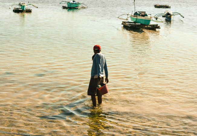 Back view of fisherman holding a wading bucket in water near boats