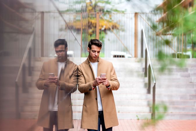 Man in fall coat leaning on reflective outdoor wall while texting