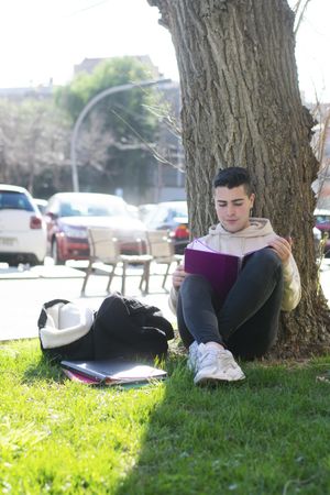 Young man leaning on a tree while reading a book