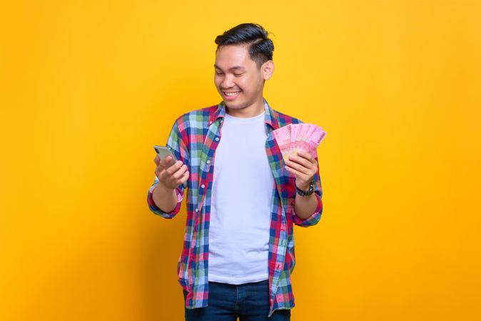 Smiling Asian man holding cash and looking down at phone screen in studio shoot