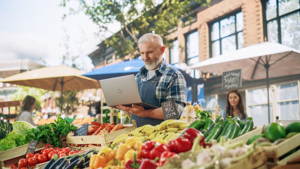 Farmer Working on a Laptop Computer, Writing Emails to Suppliers to Orders Produce for His Organic Street Vendor Stall