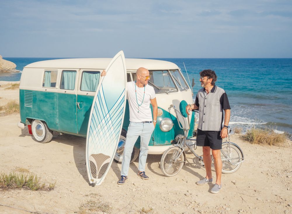 Men with surfboard and bicycle standing near car on dusty seaside