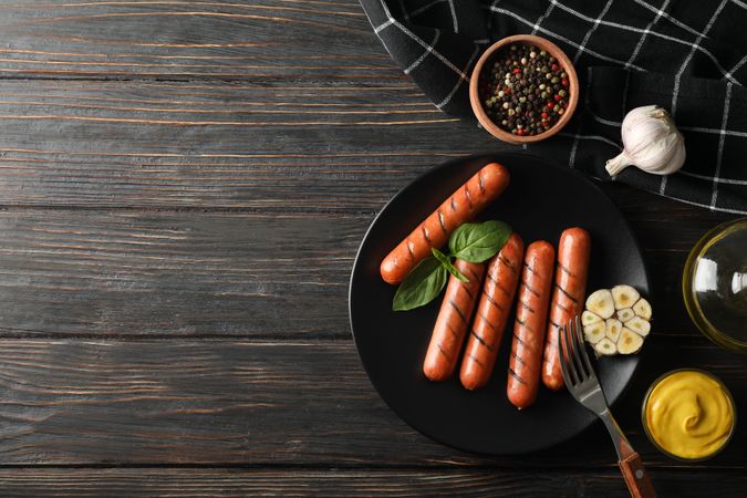 Top view of grilled sausages or hot dogs on a fork on dark ceramic plate with garlic, copy space