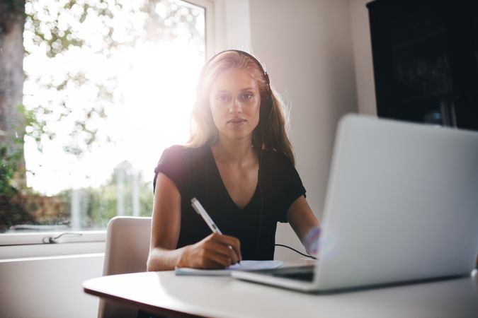 Portrait of young woman sitting in dining room with headphones writing note
