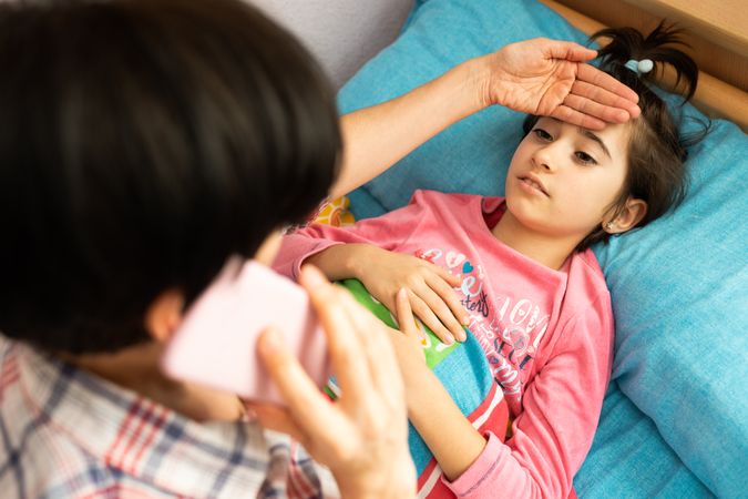 Woman on phone while checking daughter’s temperature with hand on forehead