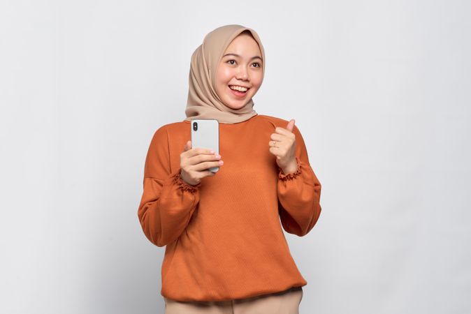 Muslim woman in headscarf and orange shirt celebrating and holding phone