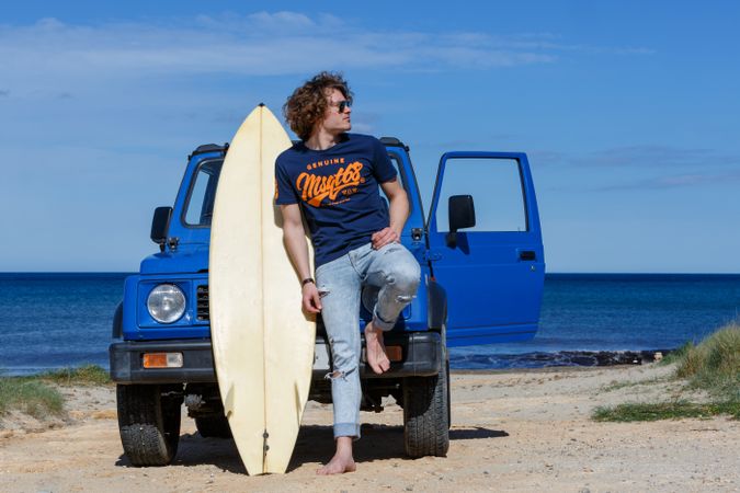 Guy with surfboard barefoot leaning on car on beach