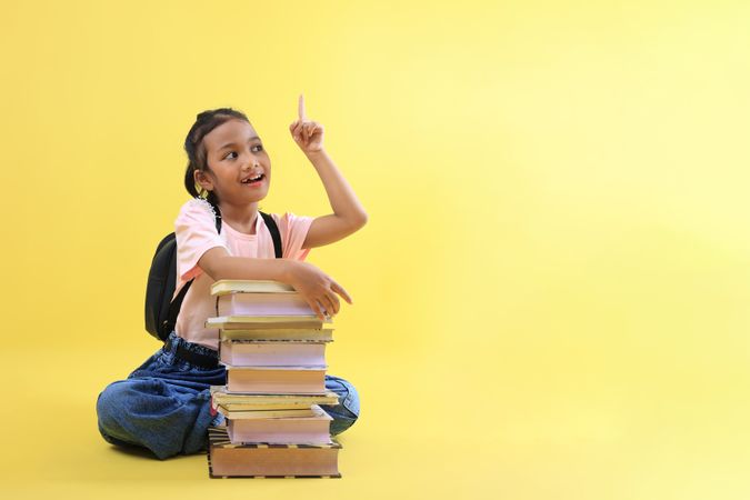 Little Asian Girl Sitting Wearing Casual Clothes and Jeans, Pointing to the Top, Her Hand on Stacked Book.