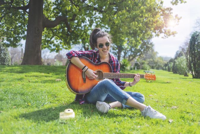 Front view of a young hipster woman wearing sunglasses, sitting on grass in a park while enjoying playing guitar